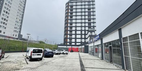 Parke taşı döşeli otopark avlusu ve park etmiş araçlar; solda çok katlı konut kulesi, sağda vitrinli tek katlı dükkân sırası