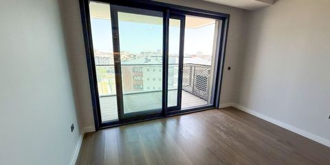 Boşaltılmış bir dairenin ahşap zeminli odası; geniş sürme cam kapının ardında balkon ve çevredeki çatılar