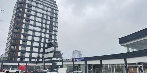 Kapalı havada çok katlı bir konut kulesi; önünde otopark ve tek katlı dükkân sırası